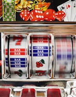 A close-up view of a slot machine with vibrant colors and various gambling symbols such as dice, coins, and playing cards. The reels display a combination of symbols, including bars, cherries, and the number seven. The chrome exterior and colorful details convey the theme of casino and gambling.