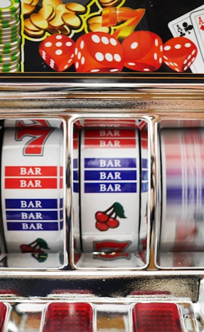 A close-up view of a slot machine with vibrant colors and various gambling symbols such as dice, coins, and playing cards. The reels display a combination of symbols, including bars, cherries, and the number seven. The chrome exterior and colorful details convey the theme of casino and gambling.