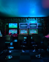 Dimly lit gaming arcade featuring a variety of brightly illuminated slot machines and video gaming screens. Neon signs and colorful displays create a vibrant atmosphere. The arrangement of gaming machines suggests an inviting space for entertainment, with chairs positioned for players.