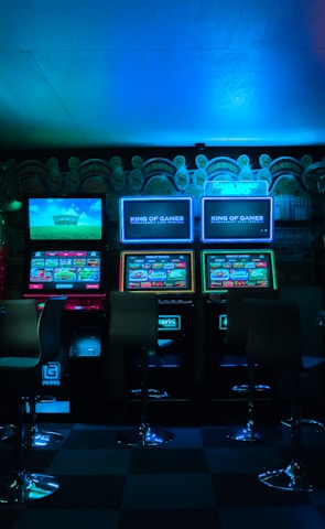 Dimly lit gaming arcade featuring a variety of brightly illuminated slot machines and video gaming screens. Neon signs and colorful displays create a vibrant atmosphere. The arrangement of gaming machines suggests an inviting space for entertainment, with chairs positioned for players.
