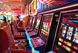 A brightly lit casino interior featuring a row of colorful slot machines. The neon lights create a vibrant atmosphere, with patterned carpeting adding to the decor. Stools are aligned neatly in front of each machine.