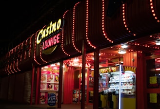 A brightly lit casino lounge entrance adorned with neon lights. The exterior features colorful signage including the words 'Casino' and 'Lounge' in vivid colors, with additional lights framing the building. Inside, a glimpse of a claw machine filled with plush toys and arcade games is visible. The area exudes an inviting yet bustling atmosphere typical of entertainment venues.