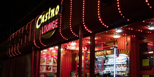 A brightly lit casino lounge entrance adorned with neon lights. The exterior features colorful signage including the words 'Casino' and 'Lounge' in vivid colors, with additional lights framing the building. Inside, a glimpse of a claw machine filled with plush toys and arcade games is visible. The area exudes an inviting yet bustling atmosphere typical of entertainment venues.