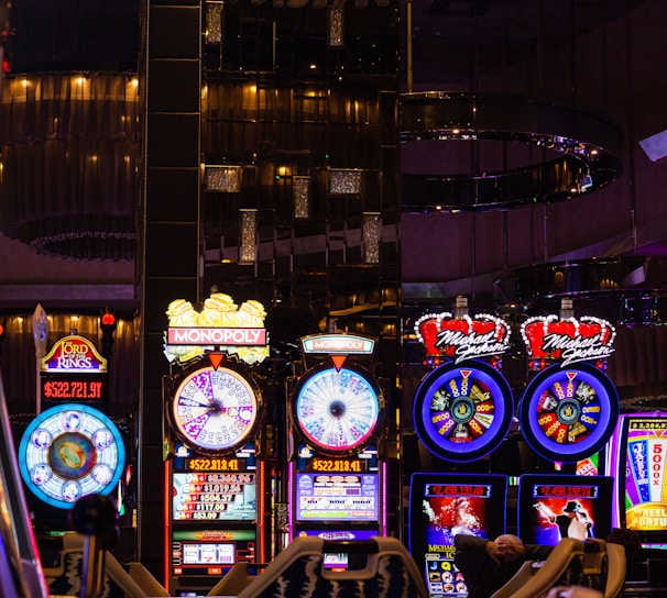 A lively casino scene featuring a row of bright, colorful slot machines with themes like Monopoly, Michael Jackson, and Lord of the Rings. The gaming machines display a variety of flashing lights and digital screens, creating an energetic and exciting atmosphere. Reflections of the lights are visible in glossy surfaces above the machines.