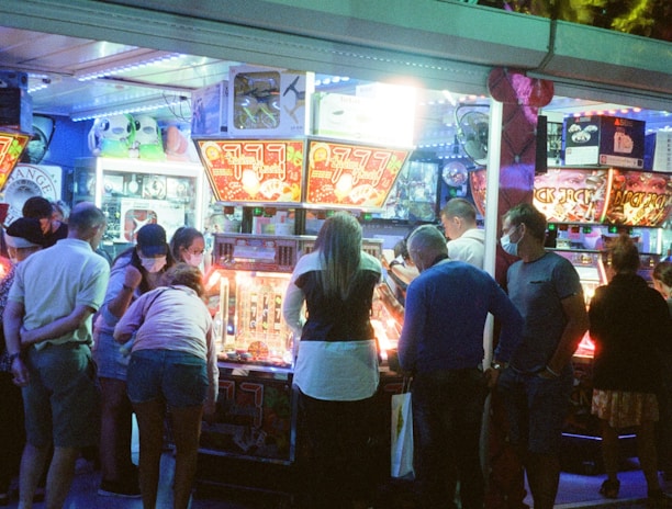 A group of people standing in front of an illuminated amusement arcade, engaging with various gaming machines. The environment is bustling with activity, and colorful lights create a vibrant atmosphere. The ceiling is decorated with large, playful images of dice and other game-related graphics, enhancing the lively mood.
