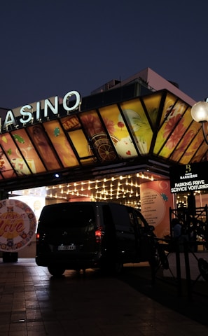 A building with a brightly illuminated entrance is seen at night. The sign above reads 'Casino' in bright letters. The facade features colorful graphics depicting various indulgent themes. Several vehicles are parked near the entrance, and people are visible in the vicinity. Tall lamp posts with spherical lights are present, casting a warm glow.