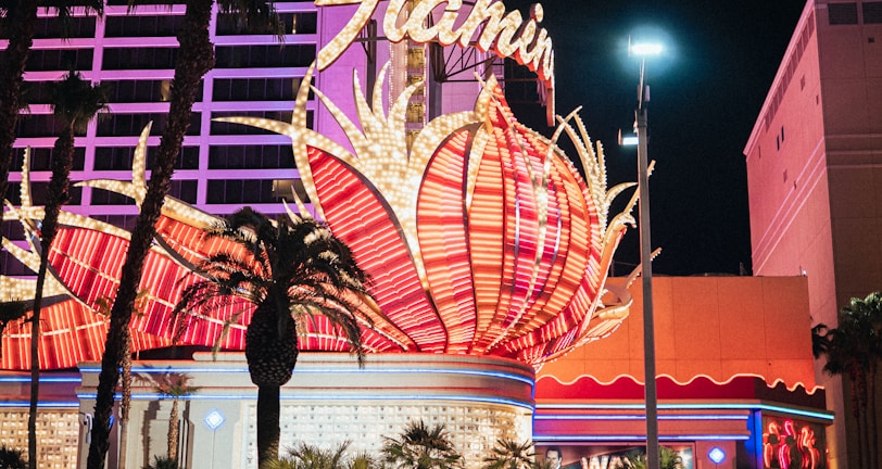 A brightly lit casino with neon lights in vibrant red, pink, and blue, featuring a large flamingo signage. Palm trees are visible in the foreground with vehicles passing by on the street at night.